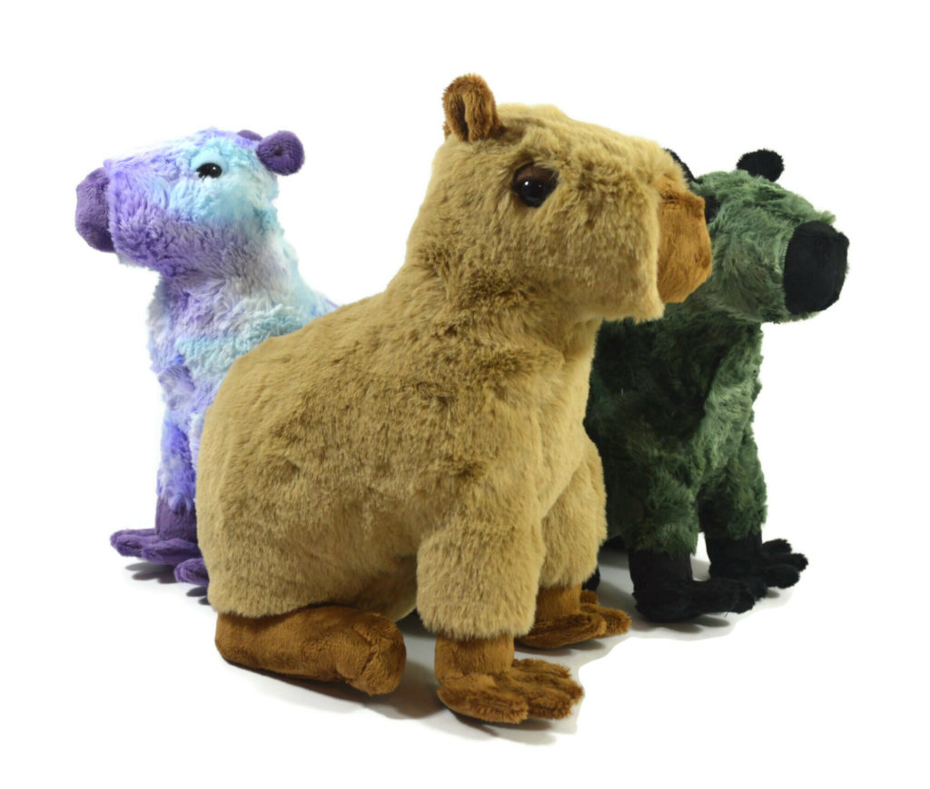 A trio of capybara plush arranged with a tan and brown one in the front, a green and black one on the right, and a blue and purple one on the left.